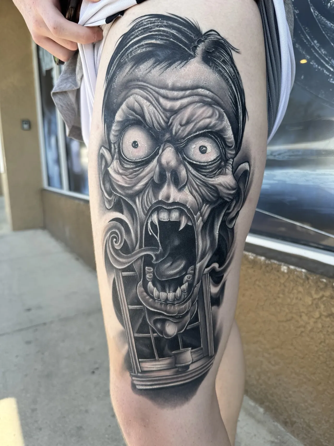 Black and grey realism tattoo of a screaming ghoul face in a window frame, on a thigh