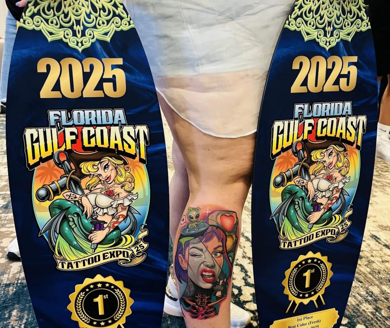 Color portrait tattoo with a small alien and a heart on a calf, between two award boards from the Florida Gulf Coast Tattoo Expo 2025